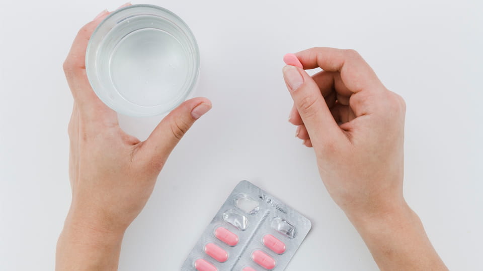 Person holding a pink pharmaceutical tablet and a glass of water, with a blister pack of tablets on the table.
