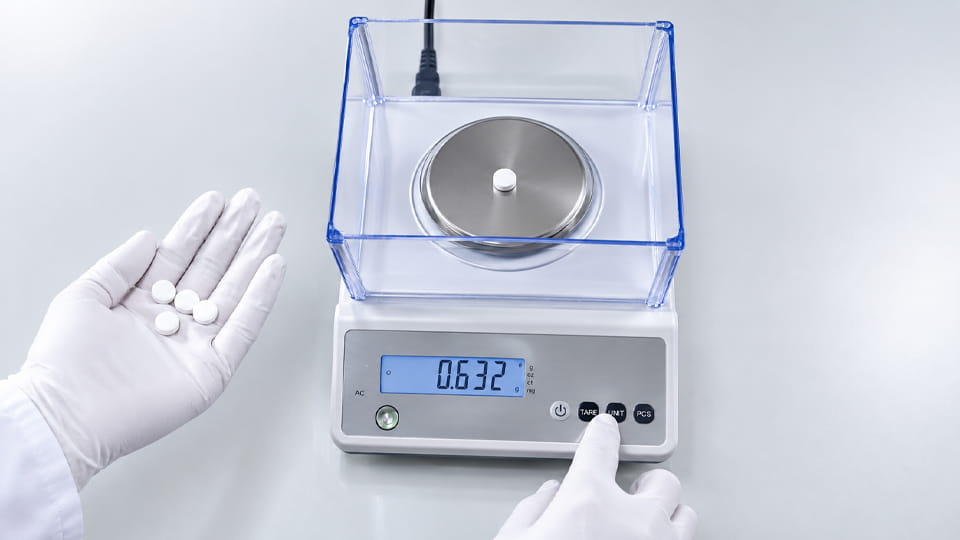 Gloved hand holding tablets beside a digital scale during tablet weight checking.
