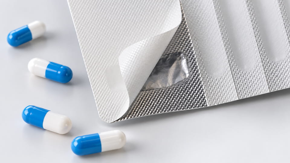 Peel-push blister packaging with a lifted foil lid exposing the silver foil layer, alongside blue-and-white capsules.