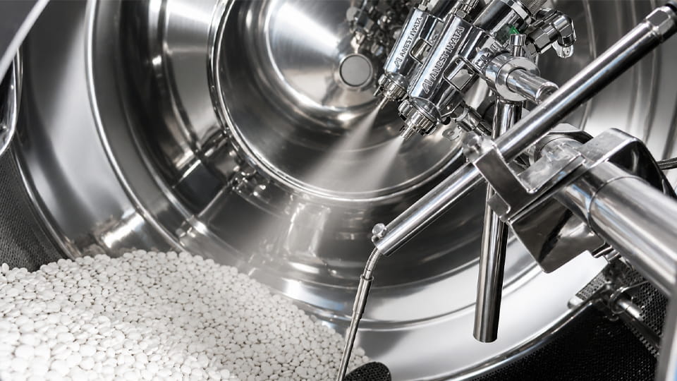 Pharmaceutical tablet coating machine with white tablets inside the coating pan and spray nozzles above the tablet bed.